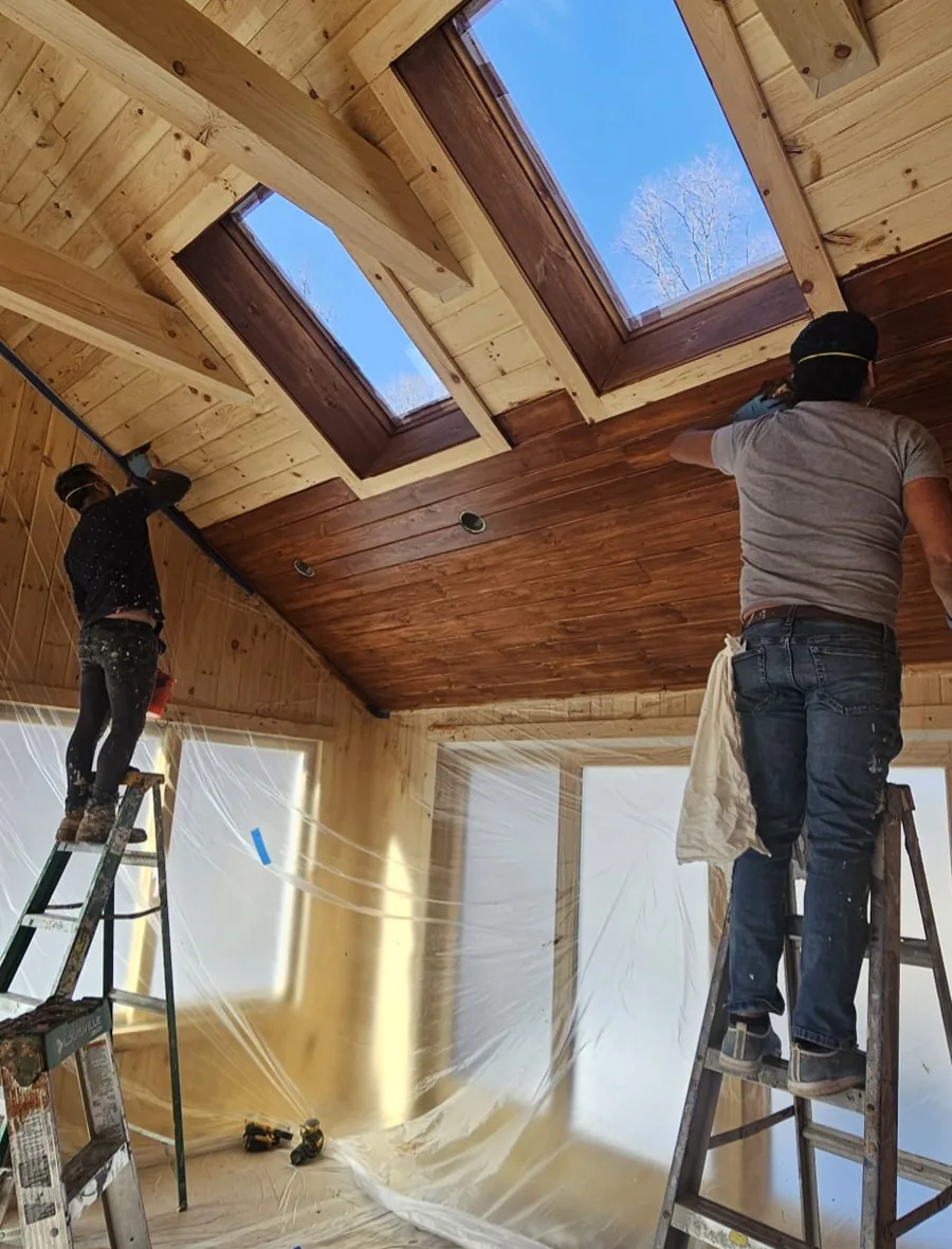 Crew staining a vaulted tongue-and-groove wood ceiling with skylights, room fully masked off